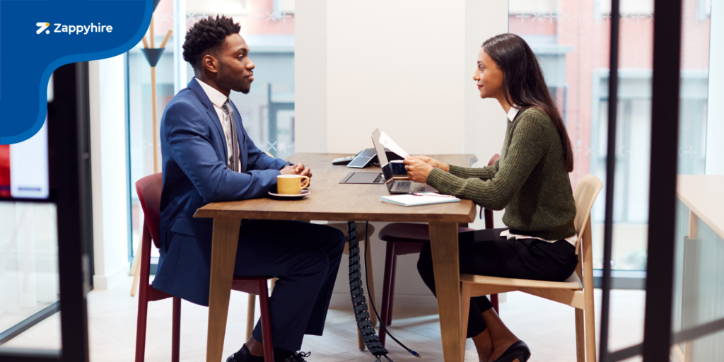 A candidate in a blue suit is being interviewed by a recruiter in a green sweater, seated across a table with laptops and documents, depicting a structured interview environment.
