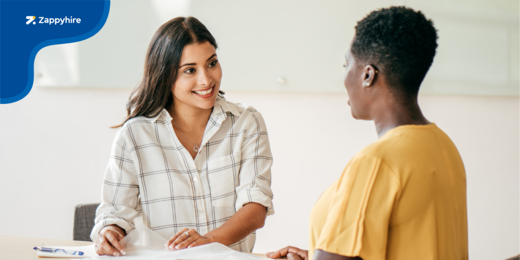 A female candidate in a checkered shirt smiles while being interviewed by a recruiter in a yellow top, showcasing a friendly and professional structured interview setting.
