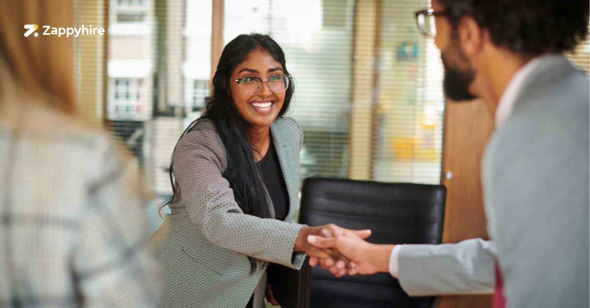 A smiling candidate, wearing glasses and a blazer, shakes hands with a recruiter in a professional setting, illustrating the beginning of a structured interview process.